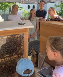 Eine Gruppe von vier Personen steht unter einem Zelt, das eine Bienenbeute zeigt. Zwei Frauen lachen, während ein Mann und eine weitere Frau lächeln. Ein Kind mit einer blauen Mütze schaut neugierig auf die Bienen. Der Hintergrund zeigt eine grüne Umgebung.