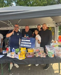 Fünf Personen stehen vor einem Stand unter einem Zelt. Sie lächeln und posieren für die Kamera. Auf dem Tisch sind verschiedene Snacks und Getränke mit einer großen Tafel, auf der Preise aufgeführt sind.