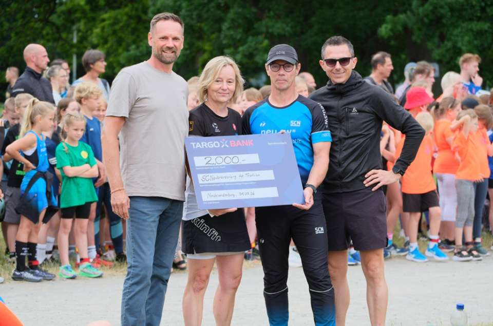 Vier Personen stehen in der Mitte eines Sportevents, in dem Hintergrund sind viele Kinder in Sportkleidung. Eine Frau hält einen großen Scheck mit der Aufschrift '2.000 €' von der TARGO BANK. Sie tragen Sportkleidung, eine Person hat eine Sonnenbrille auf. Die Umgebung ist grün und es scheint ein sonniger Tag zu sein.