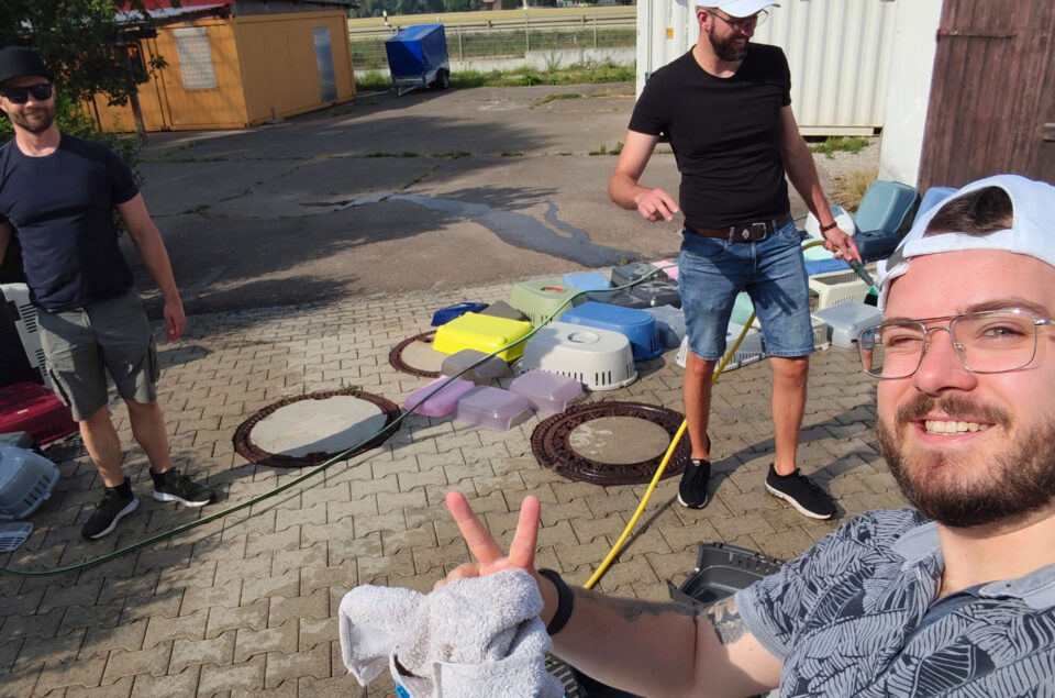 Drei Männer stehen im Freien, umgeben von bunten Behältern. Einer von ihnen macht ein Selfie und zeigt das Peace-Zeichen. Im Hintergrund sind weitere Container und ein Hänger zu sehen.