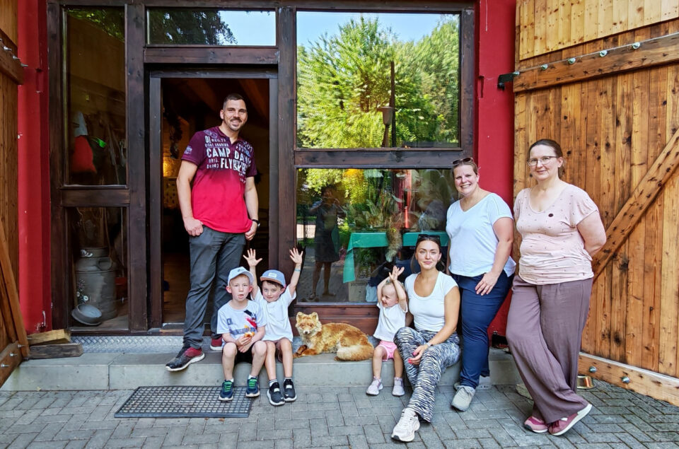 Eine Gruppe von fünf Personen steht und sitzt vor einem großen Fenster in einem Holzgebäude. Ein Mann in einem roten T-Shirt steht links, während eine Frau in einem weißen T-Shirt und eine Frau in einem rosa Oberteil rechts stehen. Vor ihnen sitzen drei Kinder und halten ihre Hände hoch, neben einem Fuchs, der auf dem Boden liegt. Das Gebäude hat eine rote Wand und eine Holztür, und im Hintergrund sind Bäume zu sehen.