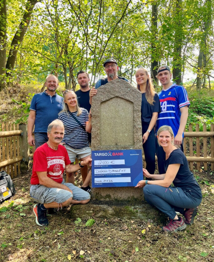 Eine Gruppe von acht Personen steht um ein Grabdenkmal im Wald. Sie halten ein großes Scheck über 2000 € von TARGO X BANK in der Hand. Die Personen sind lächelnd und tragen legere Kleidung. Im Hintergrund sind Bäume und ein Zaun sichtbar.