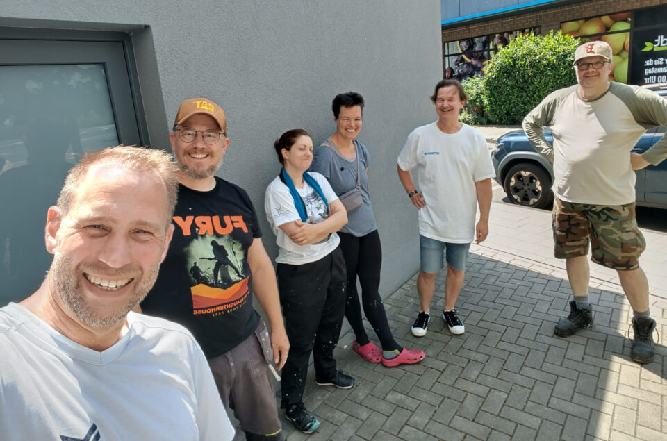 Eine Gruppe von sechs Personen steht vor einer grauen Wand. Ein Mann mit einem Bart und einem T-Shirt lächelt in die Kamera. Neben ihm steht ein weiterer Mann in einem schwarzen T-Shirt mit der Aufschrift 'FURY'. Eine Frau in einem weißen T-Shirt und einer blauen Schärpe hat die Arme verschränkt. Eine weitere Frau trägt ein graues T-Shirt und schaut leicht zur Seite. Zwei Männer stehen am Rand, einer in einem weißen T-Shirt und Shorts, der andere in einem khakifarbenen Hemd und Camouflage-Shorts. Im Hintergrund ist ein Auto und eine grüne Hecke zu sehen.