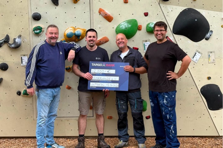 Vier Männer stehen vor einer Kletterwand und halten einen großen Scheck. Der Scheck ist auf 2.000 Euro ausgestellt und trägt die Aufschrift 'Wanderfreunde e.V.' sowie das Datum '26.09.2022'. Alle vier Männer lächeln, während sie posieren. Die Kletterwand hat verschiedene bunte Griffe in Form von Steinen.
