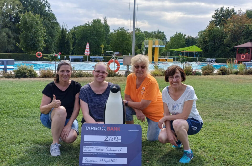 Vier Frauen stehen lächelnd vor einem Schwimmbad, während sie einen Scheck über 2000 Euro für das Freibad Gimmbheim e.V. halten. Eine Frau trägt ein oranges T-Shirt und eine andere ein gestreiftes Oberteil. Zwischen ihnen steht eine Pinguinfigur. Im Hintergrund sind Schwimmbad und grüne Bäume sichtbar.