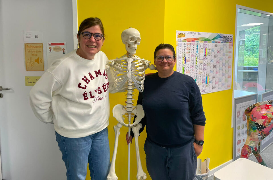 Zwei Frauen stehen neben einem Skelett in einem Raum mit gelber Wandfarbe. Eine Frau trägt einen weißen Pullover mit rotem Text, die andere ein dunkelblaues Shirt. Im Hintergrund sind ein Kalender und ein Hinweis zur Ruhe zu sehen.