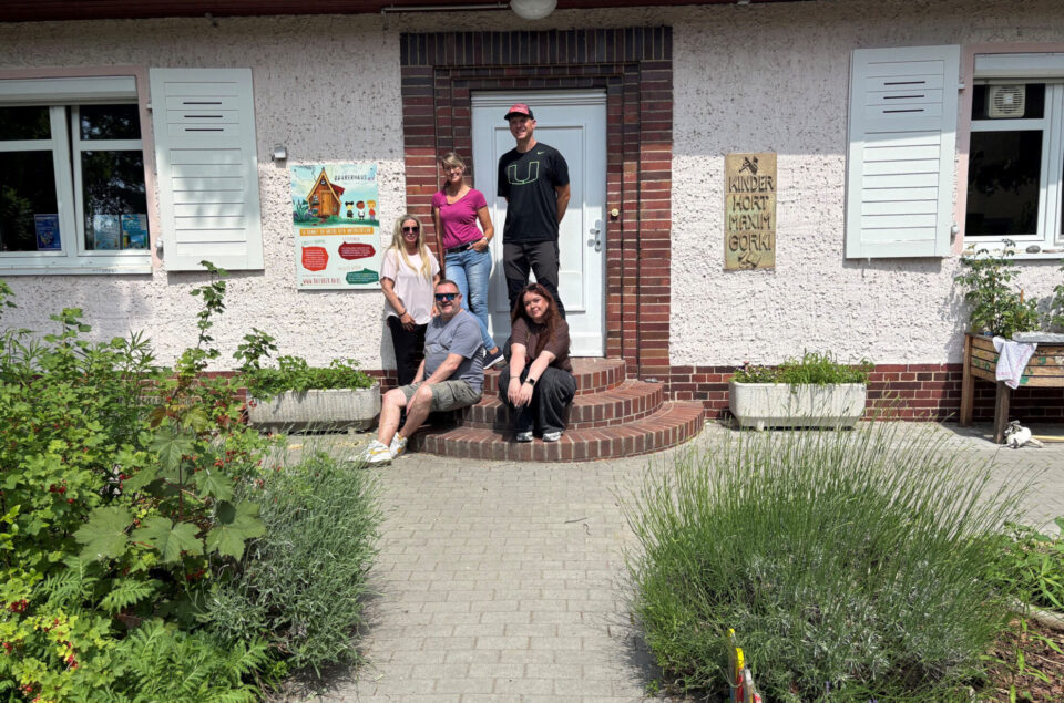 Eine Gruppe von vier Personen steht und sitzt auf den Stufen eines Hauses mit einer hellen Fassade. Im Hintergrund sind Fenster und ein Schild mit der Aufschrift "Kinder Hort Maxim Gorki" zu sehen. Die Personen sind lässig gekleidet und posieren freundlich für das Foto, während grüne Pflanzen und Blumen um die Stufen herum wachsen.