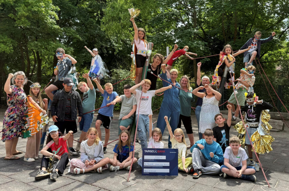 Eine Gruppe von Kindern und Erwachsenen posiert auf einem Spielplatz. Die Kinder tragen kreative Kostüme und halten eine große Scheck für 2000 Euro der TARGOBANK. Im Hintergrund sind Bäume zu sehen, und alle scheinen fröhlich und engagiert zu sein.