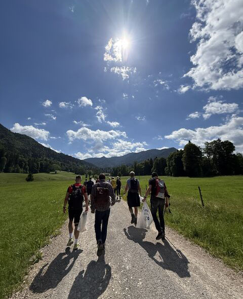 Eine Gruppe von Menschen wandert auf einem Schotterweg durch eine grüne Wiese. Der Himmel ist klar mit einigen Wolken und die Sonne scheint hell. Im Hintergrund sind Berge und Bäume sichtbar.