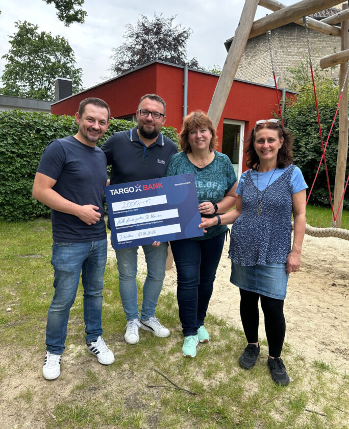 Vier Personen stehen auf einer Wiese und halten einen großen Scheck der TARGOBANK in der Höhe von 2000 Euro. Im Hintergrund sind Bäume und ein rotes Gebäude zu sehen. Die Personen lächeln und tragen legere Kleidung. Das Datum auf dem Scheck ist der 17.06.2026.