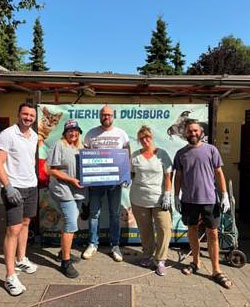 Fünf Personen stehen vor einem Tierheim in Duisburg. Sie halten ein großes Plakat und tragen Handschuhe. Der Hintergrund zeigt ein Tierheim-Schild mit Katzen und Hunden. Es ist sonnig und die Umgebung ist grün.