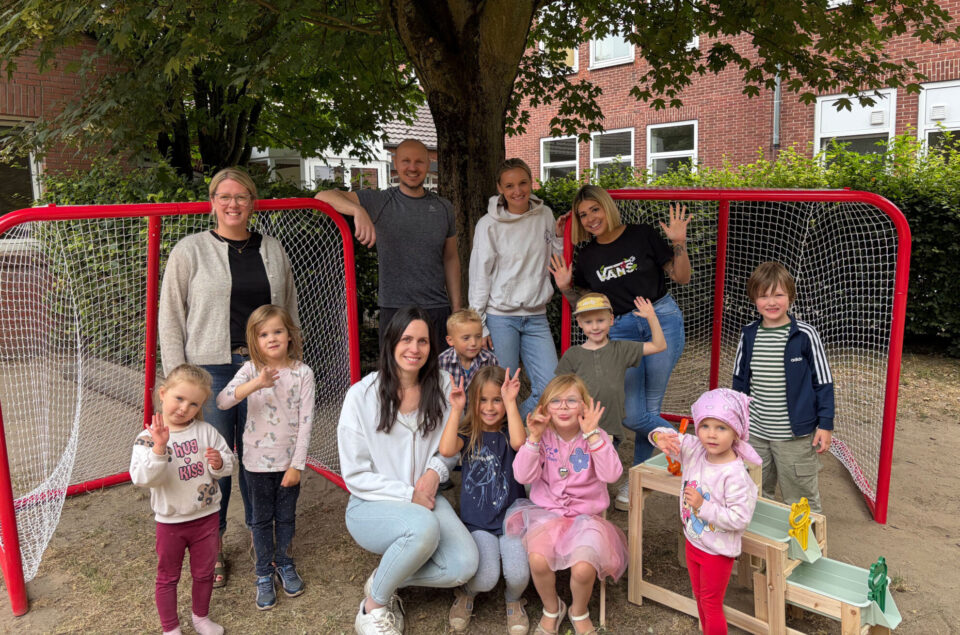 Eine Gruppe von Erwachsenen und Kindern steht unter einem Baum in einem Spielbereich. Im Hintergrund sind zwei rote Torrahmen sichtbar. Die Erwachsenen lächeln und posieren, während die Kinder verschiedene gestikulierende Posen einnehmen. Einige Kinder tragen sportliche Kleidung, während andere bunte Outfits haben. Es gibt auch eine kleine Holzkonstruktion mit Spielzeug, die im Vordergrund steht.