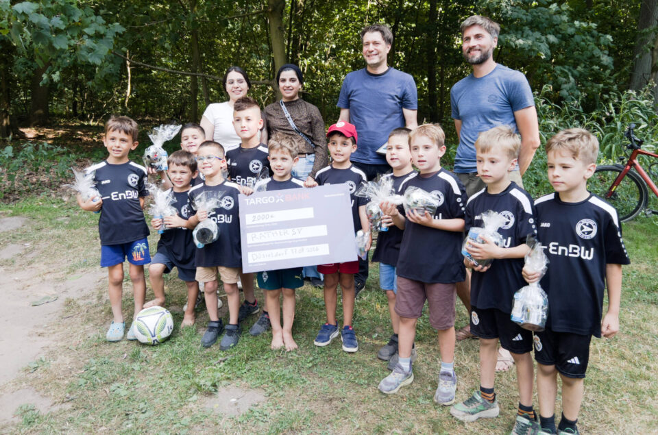 Eine Gruppe von Kindern in schwarzen Fußballtrikots mit dem Logo von EnBW steht im Freien. Sie halten kleine Geschenke in Plastiktüten und posieren vor einem großen Scheck der TARGOBANK über 2000 Euro für den RANTHER SV in Düsseldorf. Im Hintergrund sind Bäume und ein Fahrrad sichtbar.
