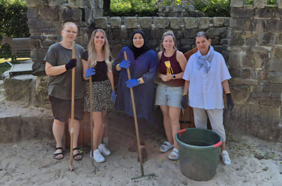 Sechs Frauen stehen in einem Sandbereich vor einer Steinmauer. Sie halten Werkzeuge in den Händen und tragen Handschuhe. Die Frauen lächeln, während sie in die Kamera schauen. Im Hintergrund sind Bäume und eine Bank sichtbar. Eine grüne Plastiktüte steht auf dem Boden.