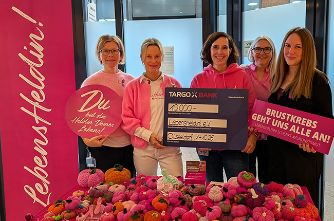 Fünf Frauen stehen lächelnd vor einem Tisch mit bunten, gehäkelten Objekten. Sie halten einen Scheck über 10.000 Euro für den Verein Lebensheldin e.V. in den Händen. Im Hintergrund sieht man eine pinke Wand mit dem Schriftzug 'LebensHeldin'. Eine der Frauen trägt ein pinkes Sweatshirt und alle sehen engagiert aus.