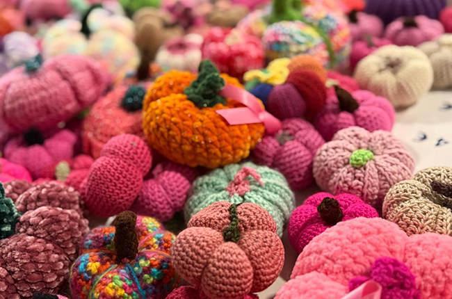 Eine Vielzahl von gehäkelten Kürbissen in verschiedenen Farben, darunter Pink, Orange, Grün und Lila, die auf einer Oberfläche verteilt sind. Die Kürbisse haben unterschiedliche Texturen und Größen, einige mit kleinen Stielen und andere mit Schleifen versehen.