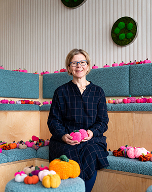 Eine Frau sitzt auf einem gepolsterten Sitzbereich mit mehreren bunten gehäkelten Kürbissen um sie herum. Sie trägt ein dunkelblaues, kariertes Kleid und hält zwei pinkfarbene Kürbisse in ihren Händen. Im Hintergrund sind weitere Kürbisse in verschiedenen Farben und Formen auf den Sitzflächen verteilt. Grüne Wanddekorationen sind ebenfalls sichtbar.