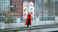 Eine Frau steht auf einem Balkon mit Glasgeländer und trägt einen roten Mantel. Im Hintergrund sind moderne und historische Gebäude sichtbar, darunter ein rotes Ziegelgebäude und ein weißes Gebäude mit roten Akzenten. Der Boden ist aus Holz und es gibt einige mit Aufklebern bedeckte Boxen.