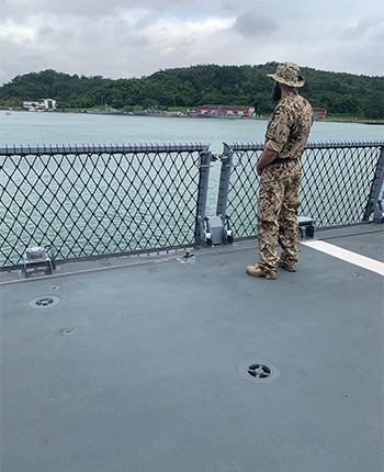 Ein Soldat in militärischer Tarnkleidung steht auf dem Deck eines Schiffes und blickt über das Wasser. Im Hintergrund sind grüne Hügel und eine kleine Siedlung sichtbar.