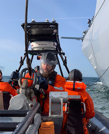 Ein Mann in orangefarbener Rettungsbekleidung steuert ein kleines Boot auf dem Wasser. Er trägt einen Helm und eine Schutzbrille. Im Hintergrund ist ein großes Schiff zu sehen. Das Wetter ist klar mit wenigen Wolken am Himmel.