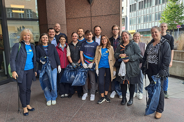 Eine Gruppe von 15 Personen posiert vor einem Gebäude. Sie tragen Handschuhe und halten Müllsäcke. Einige tragen T-Shirts mit dem Logo der Weltbank. Die Umgebung zeigt modernistische Architektur und städtische Elemente. Die Gruppe lächelt und wirkt engagiert.