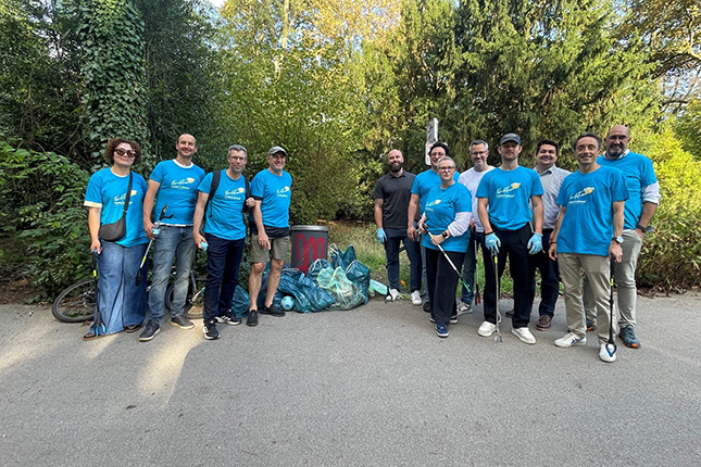 Eine Gruppe von Menschen in blauen T-Shirts steht auf einem Weg in der Natur. Sie lächeln und posieren für ein Foto. Im Hintergrund sind Bäume und Sträucher sichtbar. Vor ihnen liegen mehrere Müllsäcke, die anscheinend bei einer Aufräumaktion gesammelt wurden. Es gibt auch eine Mülltonne im Bild.