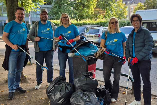 Eine Gruppe von fünf Personen steht in einem Park. Alle tragen blaue T-Shirts mit dem Logo 'Earth Day'. Sie halten Greifzangen und stehen neben einem Mülleimer, der voll mit Abfällen ist. Um sie herum liegen schwarze Müllsäcke. Im Hintergrund sind Bäume und parkende Autos sichtbar.
