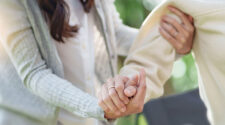 Ein Nahaufnahme von zwei Händen, die sich halten. Eine Hand trägt einen Ring und die andere Hand greift sanft eine Schulter. Im Hintergrund sind unscharfe grüne Blätter zu sehen, was eine natürliche Umgebung schafft.