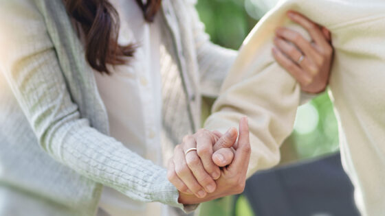 Ein Nahaufnahme von zwei Händen, die sich halten. Eine Hand trägt einen Ring und die andere Hand greift sanft eine Schulter. Im Hintergrund sind unscharfe grüne Blätter zu sehen, was eine natürliche Umgebung schafft.