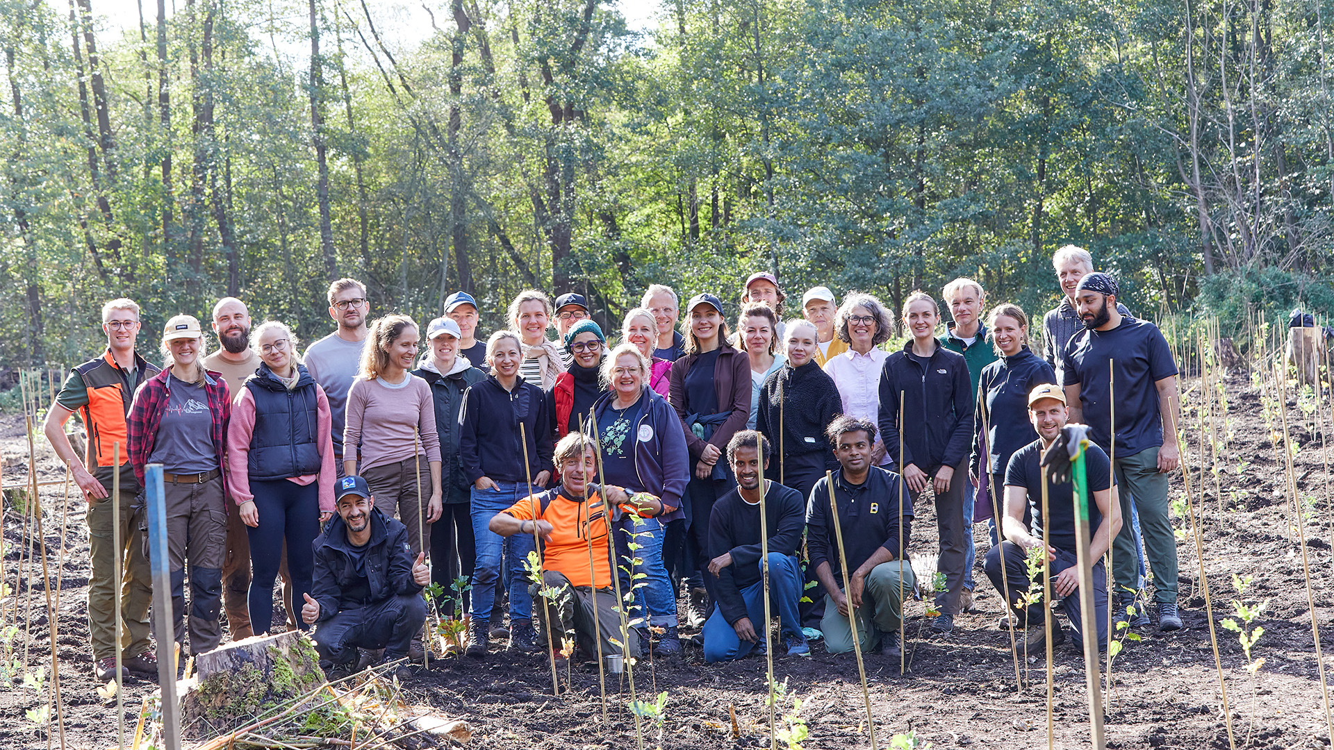 Eine Gruppe von etwa 30 Personen steht in einem Waldgebiet. Die Teilnehmer tragen verschiedene Arbeitskleidung und lächeln. Im Hintergrund sind Bäume und eine unebene Erdoberfläche mit frisch gepflanzten Setzlingen zu sehen.