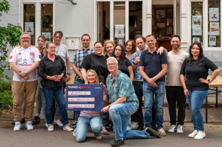 Eine Gruppe von 16 Personen steht und sitzt vor einem Gebäude. Eine Person hält einen großen Scheck der TARGOBANK über 2.000 Euro für das Café Grenzenlos e.V. in Düsseldorf. Die Gruppe wirkt freundlich und lächelt in die Kamera. Im Hintergrund sind Plakate sichtbar.