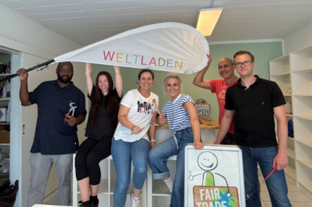 Eine Gruppe von sechs Personen steht in einem Raum mit Regalen. In der Mitte hält eine Frau ein großes Schild mit der Aufschrift 'WELTladen'. Andere Personen halten Werkzeuge oder zeigen auf ein Schild mit der Aufschrift 'FAIR TRADE'. Die Atmosphäre wirkt freundlich und einladend.