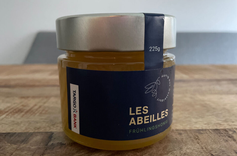 Ein Glas Honig mit einem silbernen Deckel. Die Etikette zeigt "LES ABEILLES" und "FRÜHLINGSHONIG" sowie das Logo der "TARGOBANK". Das Glas hat ein Volumen von 225g.