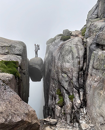 Eine Person steht auf einem schmalen Felsen, umgeben von steilen, grauen Klippen und Nebel. Die Szenerie wirkt dramatisch und abgelegen.
