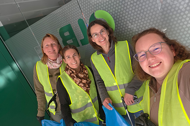 Vier Frauen stehen in einem Raum mit einem grünen Logo im Hintergrund. Sie tragen leuchtend gelbe Westen und lächeln. Zwei halten blaue Taschen.