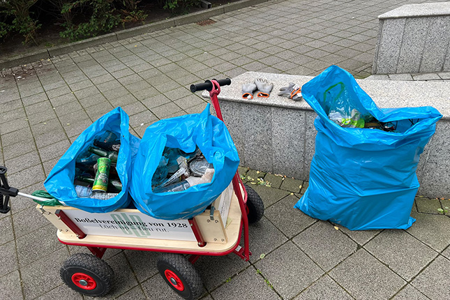 Ein roter Wagen steht auf einem gepflasterten Boden, beladen mit zwei blauen Säcken voller Müll, darunter Dosen und Flaschen. Neben dem Wagen liegen zwei Arbeitshandschuhe und ein weiterer blauer Sack.