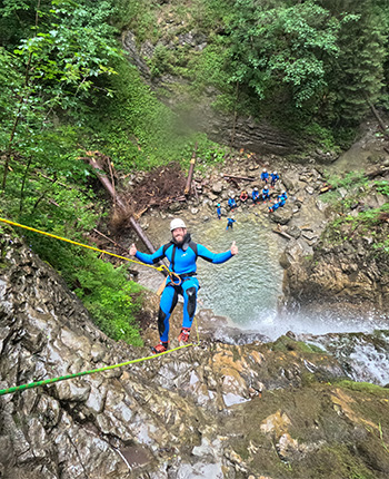 Ein Mann in einem blauen Anzug und einem Helm klettert an einem Wasserfall hinunter. Er lächelt und zeigt mit Daumen nach oben. Unten im Wasser sind Gruppen von Menschen in blauen Anzügen sichtbar.