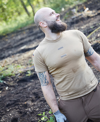Ein bärtiger Mann mit Glatze trägt ein beiges T-Shirt und braune Hosen. Er steht in einer bewaldeten Umgebung, lächelt und schaut nach oben. Seine Arme sind tätowiert, und er hat schmutzige Hände, die auf seine Arbeit hinweisen.