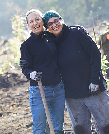 Zwei Frauen posieren fröhlich in einem Garten. Die erste Frau trägt einen schwarzen Pullover und Jeans, während die zweite Frau einen schwarzen Rollkragenpullover und eine grüne Strickmütze trägt. Beide lächeln und umarmen sich, während eine Schaufel in der Hand der ersten Frau gehalten wird. Im Hintergrund sind Pflanzen und ein verschwommener Gartenbereich sichtbar.