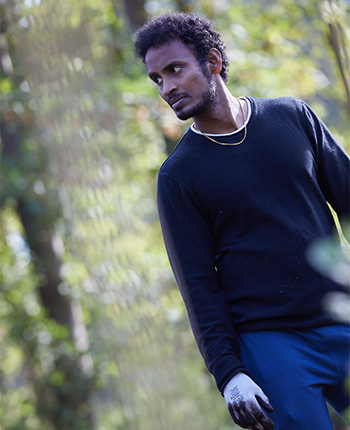 Ein Mann mit lockigem Haar trägt einen schwarzen Pullover und eine goldene Kette. Er steht in einem grünen Umfeld und schaut nachdenklich zur Seite.