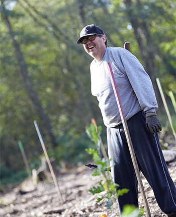 Ein Mann steht auf einem Feld, lächelt und trägt einen grauen Pullover und eine schwarzes Cap. Er hält eine lange Stange und trägt Handschuhe. Im Hintergrund sind Bäume und einige Pflanzen zu sehen.