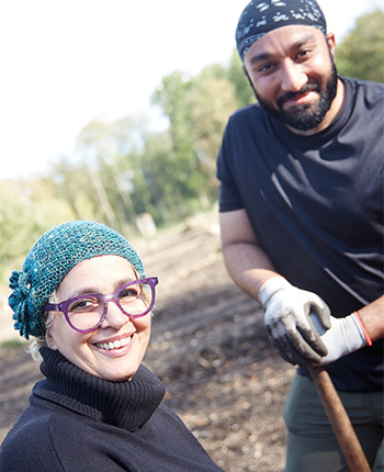 Zwei Personen arbeiten im Freien auf einem Feld. Eine Frau mit einer grünen Mütze und großen, runden Brillen lächelt in die Kamera. Ein Mann mit einem Bandana und einem Schaufelgerät steht neben ihr und schaut ebenfalls freundlich. Im Hintergrund sind Bäume und eine sonnige Landschaft sichtbar.