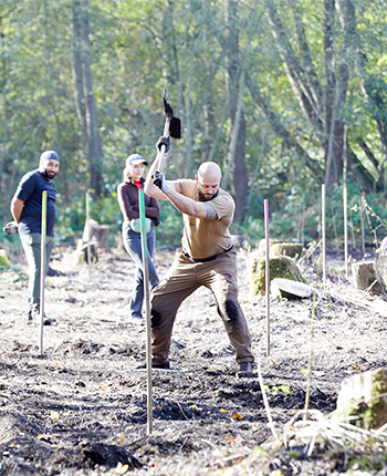 Drei Personen arbeiten im Freien in einem Waldgebiet. Eine Person, die einen Spaten hält, steht im Vordergrund und hebt ihn zum Schlagen. Im Hintergrund sind zwei weitere Personen sichtbar, eine Frau in einem grünen Oberteil und ein Mann in einem grauen T-Shirt. Rund um sie stehen Holzpfähle, die in den Boden gesteckt sind, und die Umgebung ist von Bäumen umgeben.