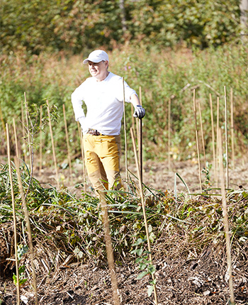 Ein Mann steht in einem Feld mit Gemüse, trägt ein weißes Oberteil und gelbe Hosen. Er hat eine Schaufel und schaut nachdenklich. Rund um ihn sind Pflanzen und Gartenstützen.