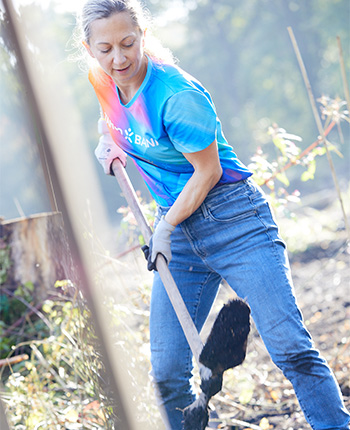 Eine Frau mit langen, blonden Haaren gräbt mit einer Schaufel in einem Waldgebiet. Sie trägt ein blaues T-Shirt mit einem Farbverlauf und Jeans. Die Umgebung ist grün und sonnig, mit unscharfen Pflanzen im Vordergrund.