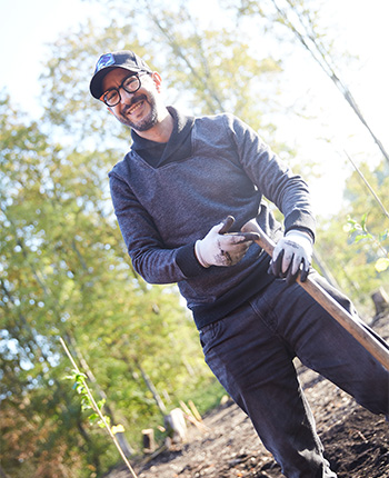 Ein Mann steht in einem Garten und lächelt. Er trägt einen grauen Pullover, schwarze Jeans und Handschuhe. In seiner Hand hält er eine Gartenschaufel. Im Hintergrund sind Bäume und Sonnenlichteinfall sichtbar.