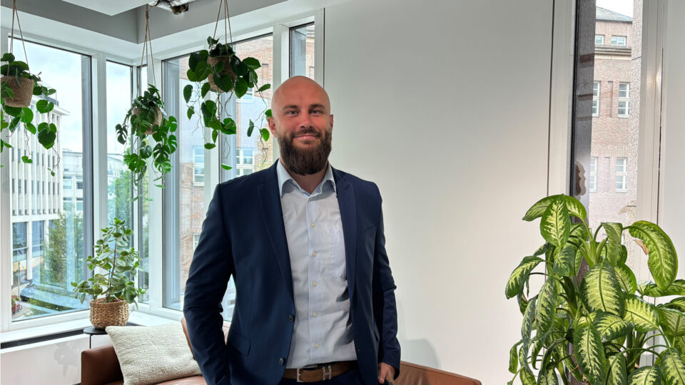 Ein Mann mit Bart steht in einem Büro mit großen Fenstern. Er trägt einen dunklen Anzug und ein helles Hemd. Im Hintergrund sind hängende Pflanzen und ein grüner Topfpflanze sichtbar.