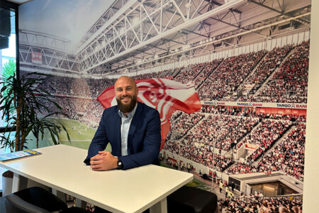 Ein Mann mit Bart und Glatze sitzt an einem Tisch in einem modernen Raum. Im Hintergrund ist eine Wand mit einem großen Bild eines Fußballstadions und jubelnden Fans. Die Atmosphäre ist sportlich und einladend.