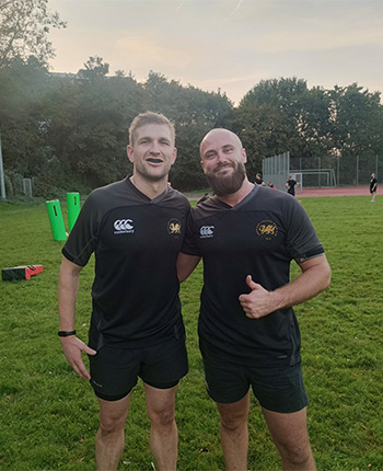 Zwei Männer stehen im Freien auf einem Sportplatz. Beide tragen schwarze Sporttrikots mit kurzen Ärmeln und Shorts. Der Mann links hat blondes Haar und lächelt, während der Mann rechts einen Bart hat und mit dem Daumen nach oben zeigt. Im Hintergrund sind grüne Trainingsgeräte und ein Sportfeld sichtbar.