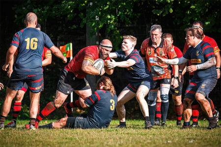 Ein Rugby-Spiel mit mehreren Spielern in actionreichen Posen. Ein Spieler in rotem Trikot mit der Nummer 3 wird von einem anderen Spieler festgehalten, während er den Ball hält. Im Hintergrund sind weitere Spieler in blau-rot gestreiften Trikots zu sehen, die das Geschehen beobachten. Die Umgebung ist grün mit Bäumen und einem klaren Himmel, was auf ein sonniges Wetter hindeutet.